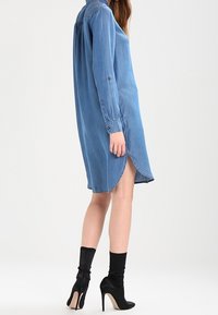 Jeansklänning i ljusblått, med knäppning fram, långa ärmar, sidoklyftor, och en avslappnad passform, kombinerad med svarta högklackade stövlar.
