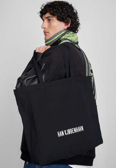 Črn platneni nahrbtnik z belim vezenim besedilom "HAN KJOBENHAVN." Ima dve trdni ročaji in preprost, strukturiran obliko.