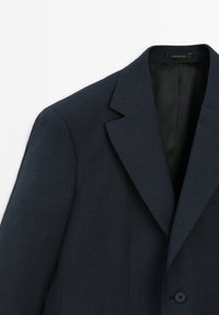Navyblauwe blazer van een gestructureerde stof, met notch-lapels en een enkele knoopsluiting. Het interieur heeft een gladde zwarte voering.
