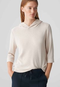 Hoodie rosa chiaro con maniche a tre quarti, tessuto testurizzato, tasca frontale e cappuccio con cordino, abbinato a pantaloni scuri.