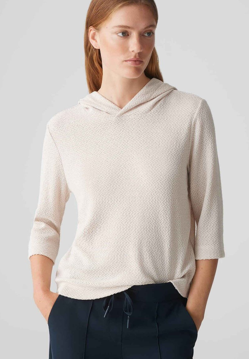 Hoodie rosa chiaro con maniche a tre quarti, tessuto testurizzato, tasca frontale e cappuccio con cordino, abbinato a pantaloni scuri.