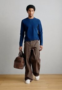 Μπλε σκούρο φούτερ, καφέ παντελόνι με φαρδύ πόδι και λευκά αθλητικά παπούτσια· κρατώντας καφέ δερμάτινη τσάντα· μινιμαλιστικός σχεδιασμός με καθαρές γραμμές.