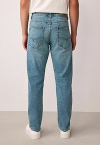 Lyseblå denimjeans med afslappet pasform, der har to baglommer og et diskret tekstureret mønster. Hvide sko ses nederst.
