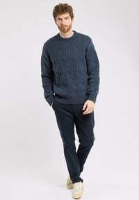 Granatowy sweter w warkocz z okrągłym dekoltem, teksturowanym wzorem i ściągaczami, w połączeniu z ciemnymi spodniami i jasnymi trampkami.