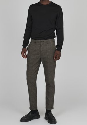 Chemise noire à manches longues associée à un pantalon marron à carreaux. Le pantalon présente une coupe ajustée et une fermeture à bouton. Des chaussures noires complètent la tenue.