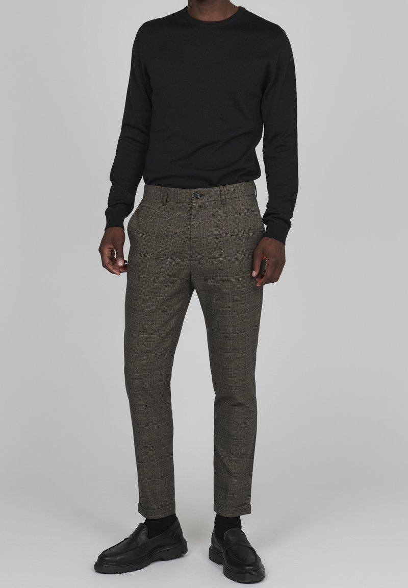 Svart långärmad skjorta som är ihopparad med bruna rutiga byxor. Byxorna har en slim fit och knappstängning. Svarta skor fulländar outfiten.