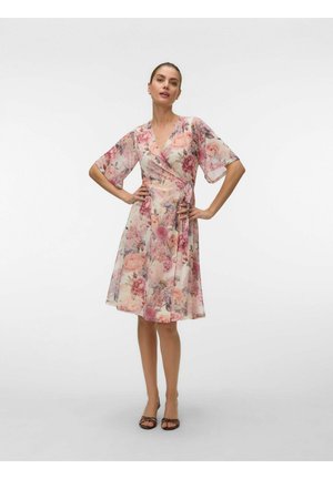 Naine kannab põlveni ulatuvat roosat ja kreemjat lillemustriga keebikleiti lühikeste varrukatega, seisab käed puusadel ja kannab musti sandaale.