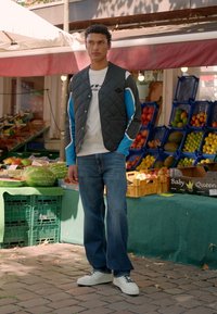 Jeune homme portant un gilet matelassé et un jean se tient les mains dans les poches près de caisses de fruits à un stand de marché en plein air.