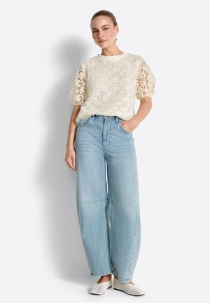 Ženska, oblečena v kremasto cvetlično bluzo z nahtraniimi rokavi, svetlo modre široke kavbojke in bele tkanine copate, stoječa z eno roko v žepu.