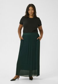 Camiseta negra de manga corta combinada con una falda larga de color verde oscuro con plisados. La falda cuenta con una cinturilla ancha y bolsillos laterales. Tacones negros.