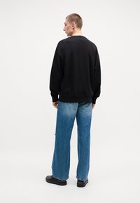 Felpa nera con vestibilità rilassata, abbinata a jeans azzurri a gamba larga e scarpe nere, vista da dietro su uno sfondo semplice.