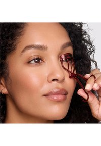 Piegaciglia rosso in uso, con una struttura in metallo, pad in silicone arrotondato e impugnatura a forbice. Il modello ha capelli ricci e un incarnato naturale.