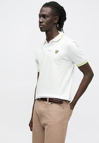 Fehér galléros póló sárga díszítéssel a gallérnál és az ujjain, kis fekete logóval. Világosbarna, texturált nadrággal viselve.