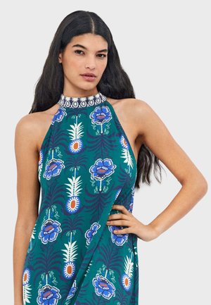 Mujer con cabello largo y oscuro, vestida con un vestido halter verde sin mangas con patrones florales azules y blancos, con una mano en la cadera.