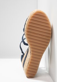 Semelle d'une sandale espadrille bleu marine avec un dessous en caoutchouc côtelé et une semelle intermédiaire en jute tressé, positionnée contre un mur d'angle blanc.