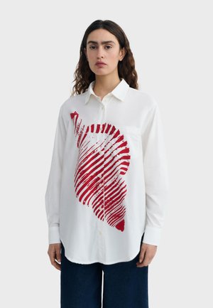 Žena s vlnitými hnedými vlasmi nosí biele košeľové tričko s abstraktným červeným pruhovaným vzorom vpredu a tmavomodré nohavice.