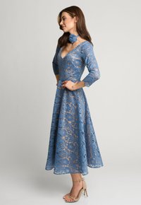 Niebieska koronkowa sukienka z dekoltem w serek, rękawami o długości trzech czwartych i kwiatowym wzorem. Zawiera pasującą ozdobę kwiatową przy szyi. Widać sandały na obcasie.