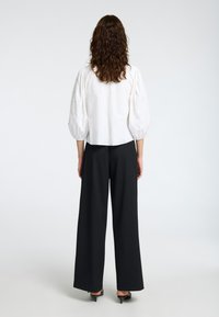 Blouse blanche à manches bouffantes avec des détails texturés, associée à un pantalon large noir taille haute et des talons noirs. Vue arrière affichée.