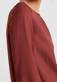 Maronin pitkähihaainen paita, jossa on pyöreä kaula-aukko ja teksturoitu kangas. Paidassa on hienovaraisia rypytyksiä olkasaumassa lisäyksinä.