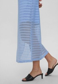Abito maxi lavorato a maglia di colore blu chiaro con righe orizzontali, caratterizzato da un design aperto. Abbinato a mule nere con tacco aperto.