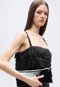 Jeune femme aux longs cheveux bruns portant un haut noir sans manches avec un design superposé en forme de pétales, tenant une pochette noire brillante.