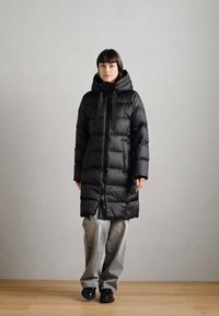 Cappotto imbottito nero con cappuccio, dotato di zip frontale, motivo a righe verticali e tasche laterali. Indossato sopra pantaloni larghi grigio chiaro.
