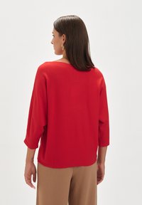 Czerwona dzianinowa bluzka z rękawami 3/4 i luźnym krojem, w zestawieniu z beżowymi spodniami o wysokim stanie. Gładka tekstura i prosty design.