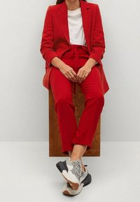 Costume rouge sur mesure avec manches longues et pantalon assorti, associé à une chemise blanche et des baskets grises. Tissu lisse, design épuré.