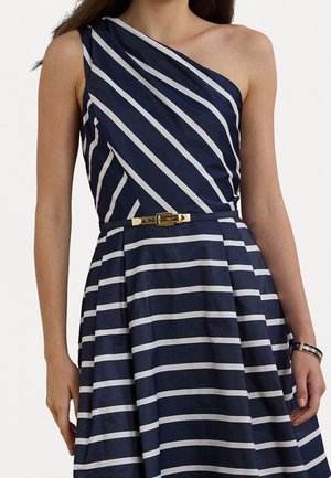Frau trägt ein marineblau-weiß gestreiftes One-Shoulder-Kleid mit einem dünnen Gürtel und passendem Armband am rechten Handgelenk.