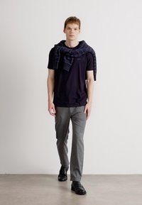 Μαύρη μπλούζα και γκρι παντελόνι, με ναυτικό μπλέ καρό πουλόβερ ριγμένο στους ώμους και μαύρα παπούτσια. Ουδέτερο φόντο.