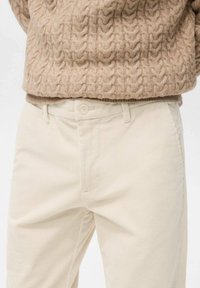Beżowy sweter dziergany z wzorem warkocza, zestawiony z jasnobeżowymi sztruksowymi spodniami z zapięciem na guziki i subtelnymi pionowymi paskami.