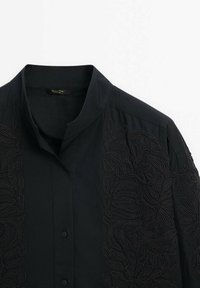 Chemise noire à boutons avec col mandarin et motifs floraux brodés ton sur ton sur les manches et le devant.