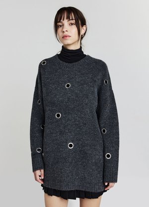 Young woman wearing a dark gray knit sweater with metal eyelet details and a black turtleneck underneath, standing against a plain background.