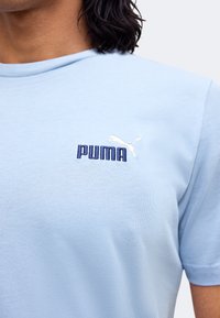 Άτομο που φοράει ένα ανοιχτό μπλε T-shirt με μικρό σκούρο μπλε λογότυπο Puma και λευκό εικονίδιο Puma στο πάνω αριστερό μέρος του στήθους.