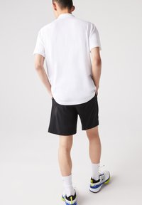 Vit kortärmad polo skjorta tillsammans med svarta shorts. Modellen har på sig vita och blå sportskor med gr öna detaljer och vita strumpor.