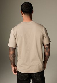 Mann mit kurzen lockigen Haaren und tätowierten Armen, der ein beiges T-Shirt und schwarze Jeans trägt, von hinten vor einem einfarbigen Hintergrund.