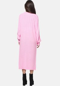 Langärmliges, pinkfarbenes Strickkleid mit gerippten Details auf dem Rücken, bis zur Mitte der Waden reichend und mit weiten Ärmeln. Dazu schwarze Knöchelestiefel.