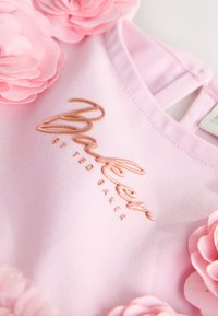 Světle růžové dětské šaty s vyšitým nápisem "Baby by Ted Baker" a měkkými růžovými látkovými květinami podél výstřihu a ramínek.