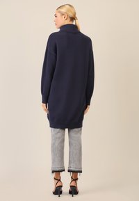 Dzianinowy sweter w kolorze granatowym z wysokim kołnierzem, luźny krój i ściągacze; w połączeniu z szarymi, krótkimi dżinsami i czarnymi sandałami na obcasie.