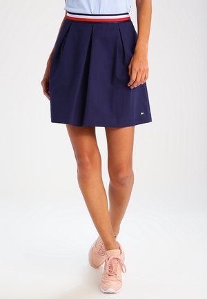 Femme portant une jupe plissée bleu marine avec une ceinture rayée rouge, blanche et bleue, ainsi que des baskets rose clair sur fond blanc.