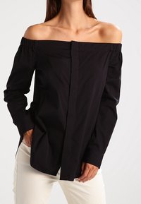 Svart off-shoulder-blus med knappfront, långa ärmar och rynkad halsringning, tillverkad av ett mjukt tyg. Vita byxor visas.