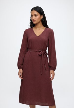 Femme aux longs cheveux foncés porte une robe bordeaux à manches longues, longueur genoux, col en V et ceinture nouée à la taille, debout devant un fond blanc uni.