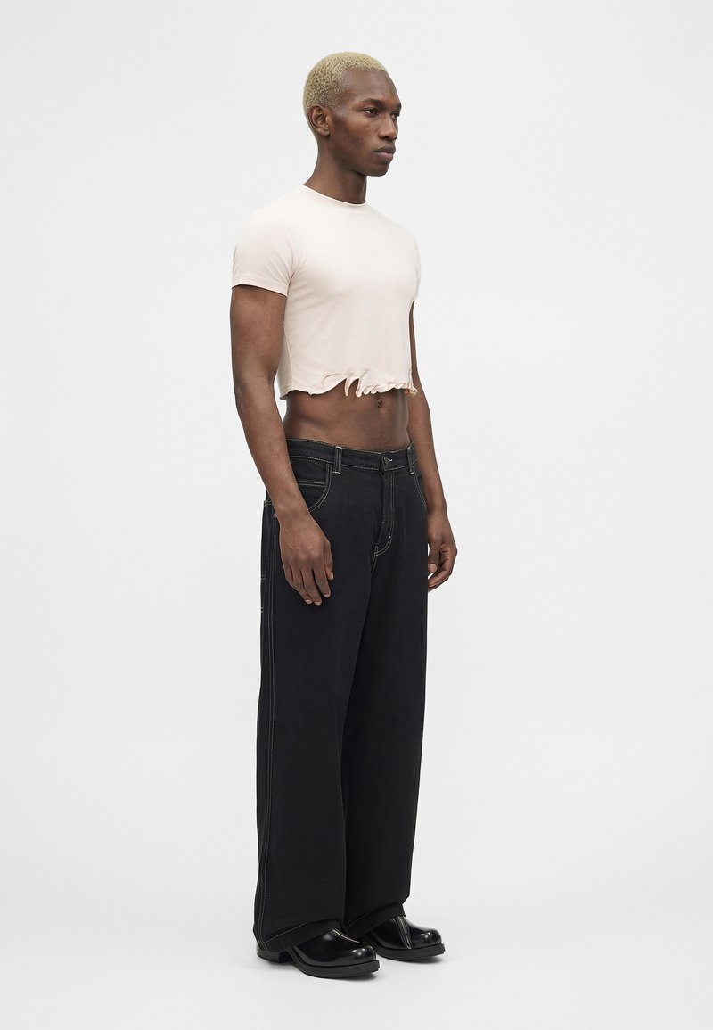 Homme aux cheveux blonds courts portant un t-shirt court rose pâle, un jean large noir et des chaussures noires, debout devant un fond blanc.