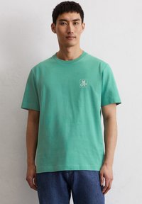 T-shirt a maniche corte, collo rotondo, in cotone verde menta con un piccolo logo bianco sul petto. Il tessuto appare morbido con una superficie liscia.