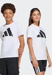 Zwei Kinder tragen weiße Baumwoll-T-Shirts mit großen schwarzen Adidas-Logos. Der Junge hat lockige Haare und das Mädchen hat lange, glatte Haare.