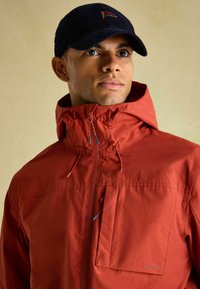 Vīrietis ar sarkanu kapuci jaku ar rāvējsl�ēdzēju un krūšu kabatu, kā arī tumši zilu cepuri ar mazu izšūtu karogu.