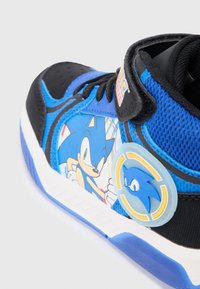 Niebieskie sneakerki z siateczkową cholewką, ozdobione kreskówkowym wzorem Sonica z Jeża oraz okrągłym logo. Czarna i biała podeszwa.