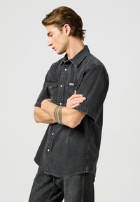 Camicia nera in denim a maniche corte con due tasche sul petto, bottoni a pressione e yoke in stile western, con una finitura leggermente testurizzata.