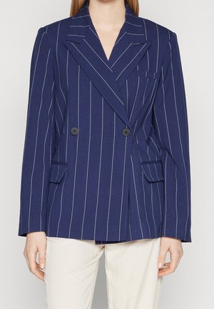 Femme portant un blazer croisé à fines rayures bleu marine avec des revers à cran et un pantalon blanc, debout devant un fond uni.