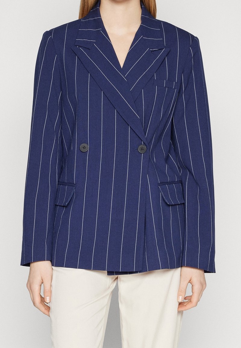 Femme portant un blazer croisé à fines rayures bleu marine avec des revers à cran et un pantalon blanc, debout devant un fond uni.